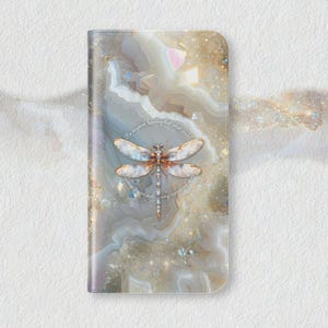 May include: A phone case with a dragonfly design. The case features a detailed dragonfly illustration in shades of white, gold, and brown, set against a background of marbled textures in neutral tones. The case has a quote in a circular pattern.