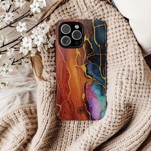 Puede incluir: Una funda de tel&eacute;fono inteligente con un dise&ntilde;o abstracto colorido. La funda presenta una mezcla de tonos c&aacute;lidos como el naranja y el marr&oacute;n, contrastados con azules y morados fr&iacute;os. L&iacute;neas doradas crean un efecto agrietado. Un peque&ntilde;o s&iacute;mbolo de br&uacute;jula es visible.