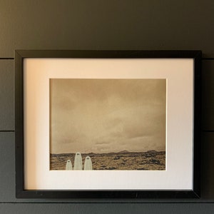 May include: A framed print of a sepia-toned landscape with three white ghosts standing in a row in the foreground.