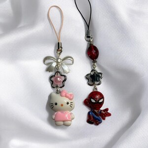 May include: Two phone charms, one with a Hello Kitty character and the other with a Spiderman character. Both charms have a silver bow and a pink and black bead.
