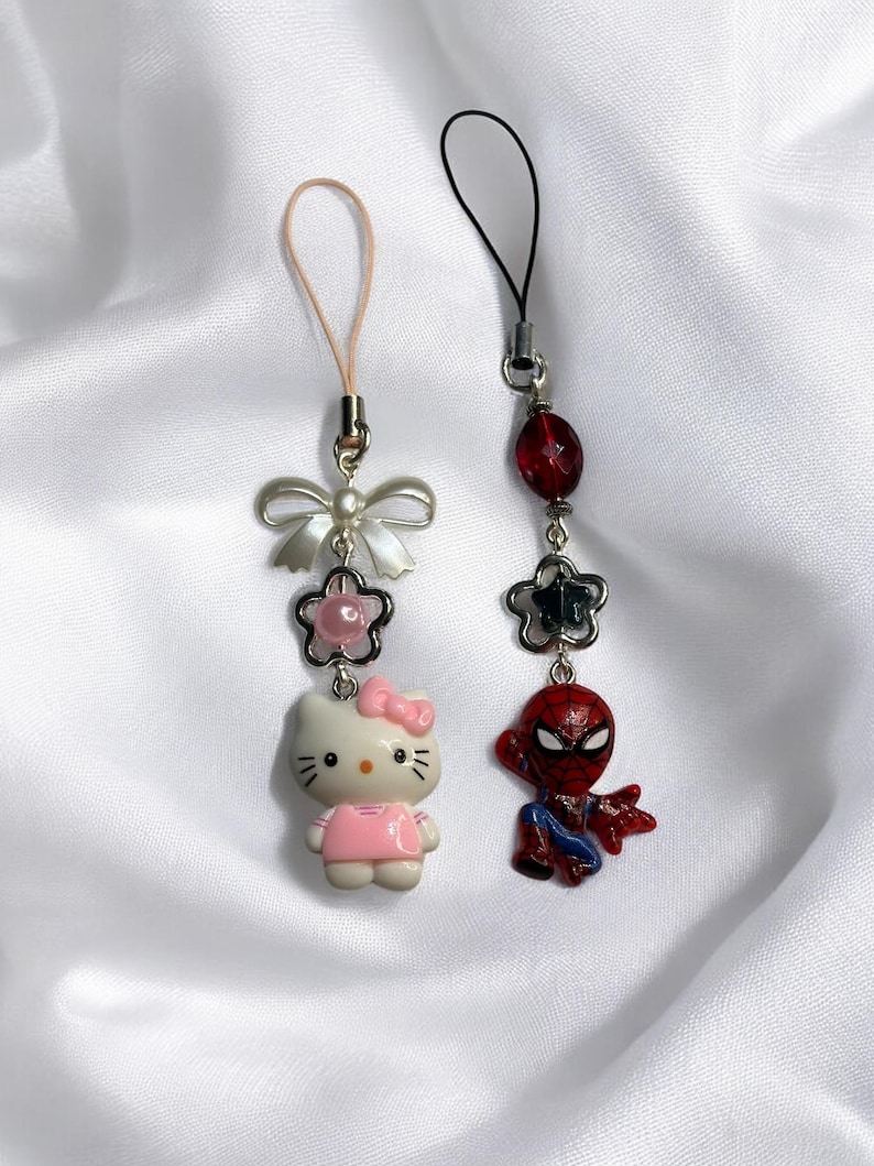 May include: Two phone charms, one with a Hello Kitty character and the other with a Spiderman character. Both charms have a silver bow and a pink and black bead.