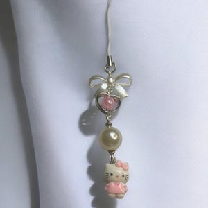 May include: A white and pink Hello Kitty charm with a pearl bead and a bow. The charm is attached to a white string.