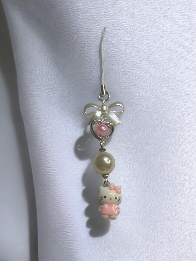 May include: A white and pink Hello Kitty charm with a pearl bead and a bow. The charm is attached to a white string.