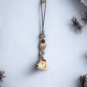 Op de afbeelding: Een witte keramische hanger van een hond met een rode kerstmanmuts, bevestigd aan een zwart touwtje met gouden en rode hartjes en heldere glazen kralen.