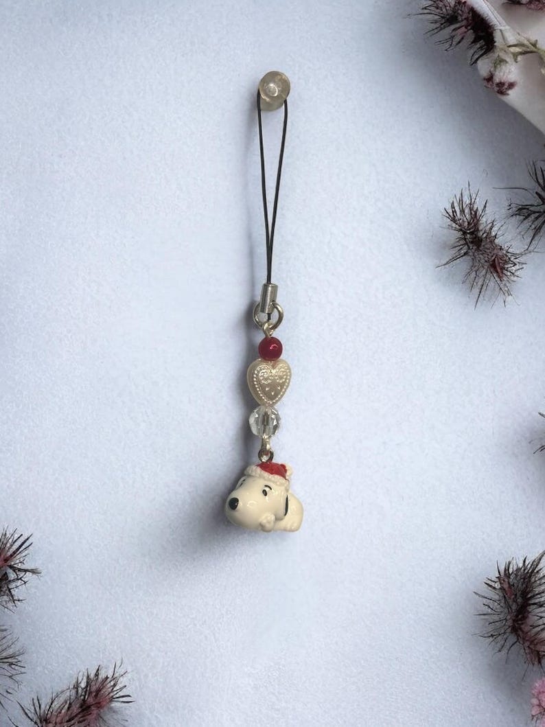 Op de afbeelding: Een witte keramische hanger van een hond met een rode kerstmanmuts, bevestigd aan een zwart touwtje met gouden en rode hartjes en heldere glazen kralen.