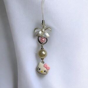 May include: A white Hello Kitty charm with a pink bow, a pearl bead, and a silver heart with a pink bead inside. The charm is attached to a clear phone strap with a silver bow.