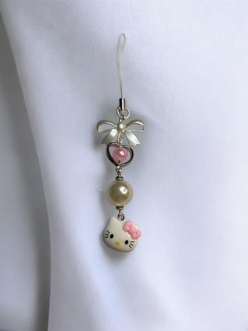 May include: A white Hello Kitty charm with a pink bow, a pearl bead, and a silver heart with a pink bead inside. The charm is attached to a clear phone strap with a silver bow.