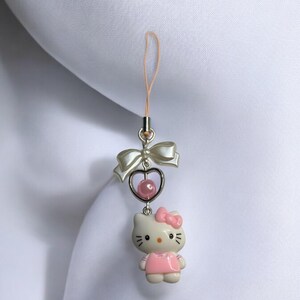 May include: A pink and white Hello Kitty charm with a heart and bow. The charm is attached to a pink string.