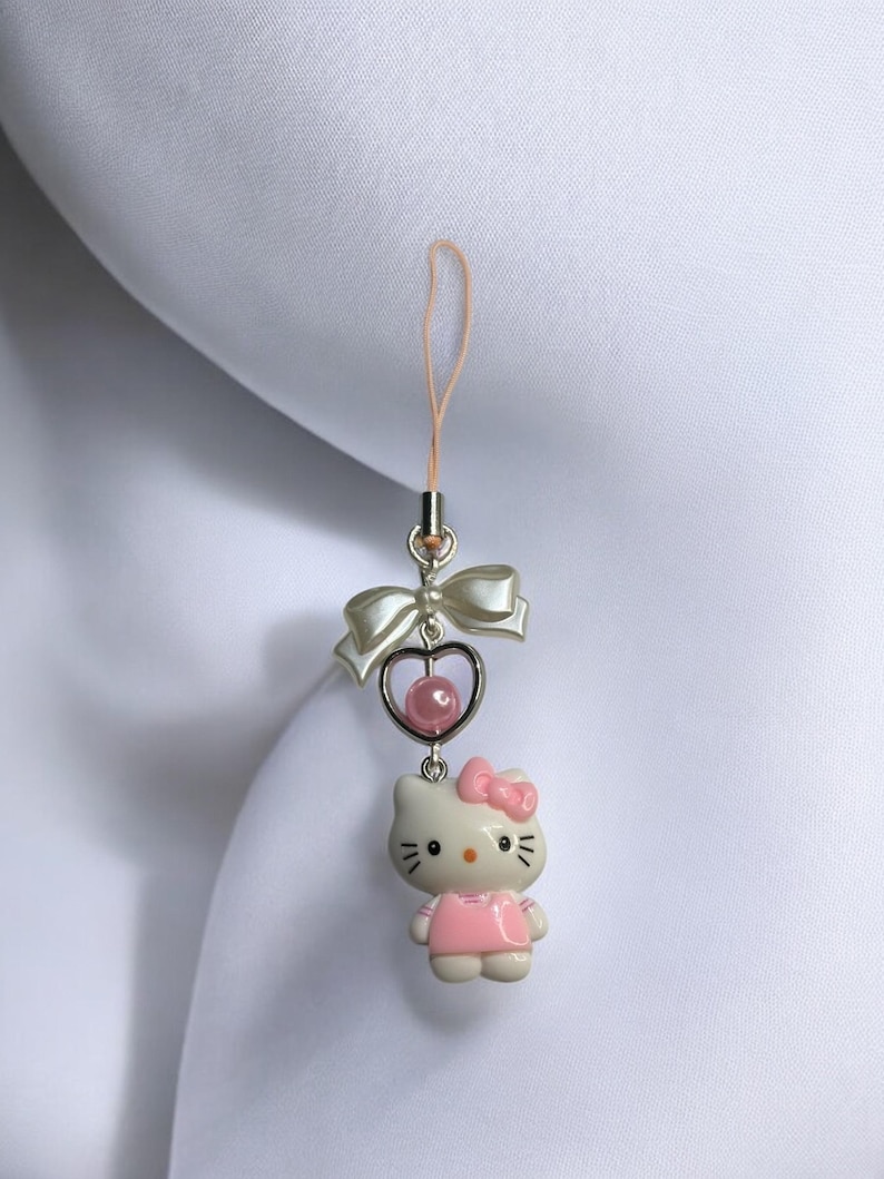 May include: A pink and white Hello Kitty charm with a heart and bow. The charm is attached to a pink string.