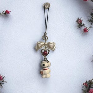 Op de afbeelding: Een witte keramische hondenhanger met een rood hart en een strik. De hanger is bevestigd aan een zwart touwtje met een zilveren clip.