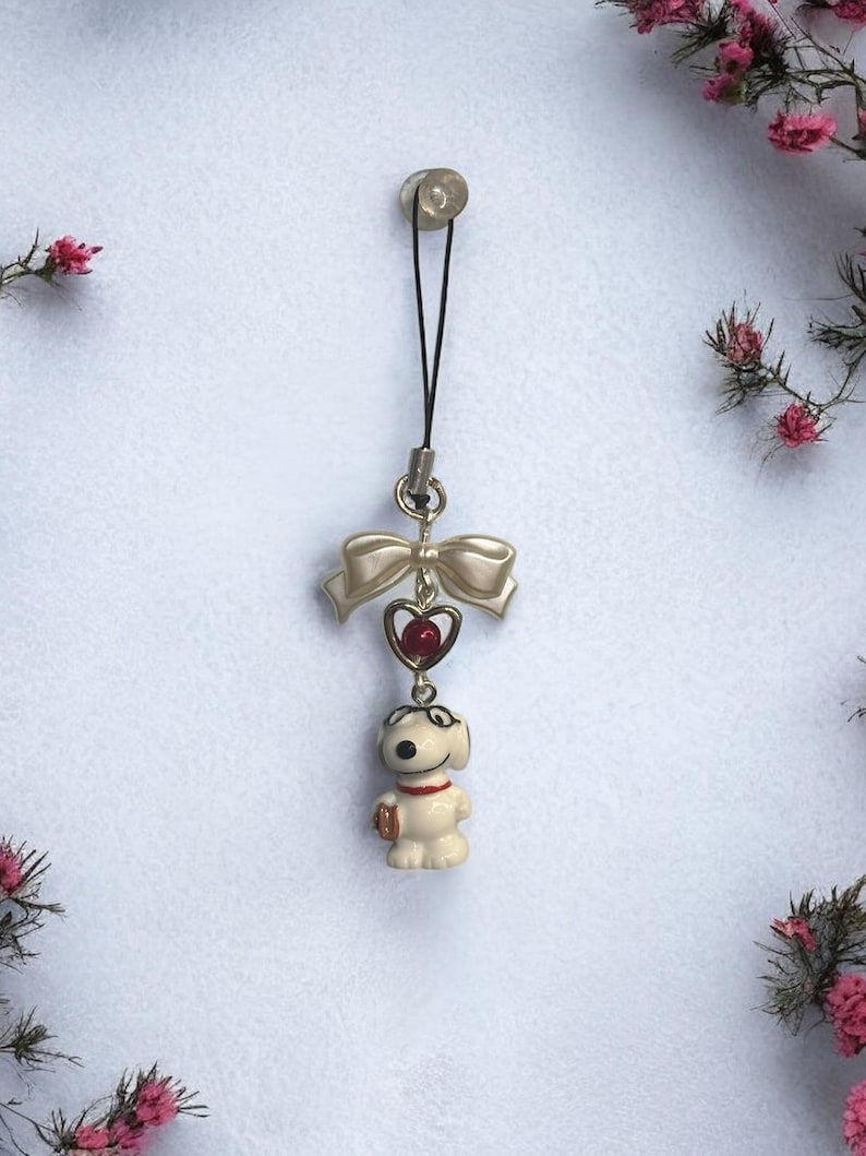 Op de afbeelding: Een witte keramische hondenhanger met een rood hart en een strik. De hanger is bevestigd aan een zwart touwtje met een zilveren clip.