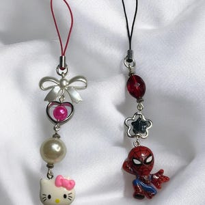 May include: Two phone charms, one with a Hello Kitty character and the other with a Spiderman character. The Hello Kitty charm has a pink bow, a white pearl, a pink heart, and a silver bow. The Spiderman charm has a red and blue Spiderman figure, a silver star, a red bead, and a black string.