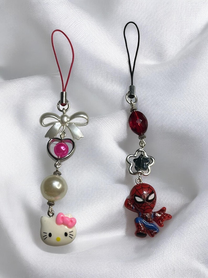 May include: Two phone charms, one with a Hello Kitty character and the other with a Spiderman character. The Hello Kitty charm has a pink bow, a white pearl, a pink heart, and a silver bow. The Spiderman charm has a red and blue Spiderman figure, a silver star, a red bead, and a black string.
