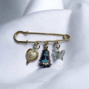 Puede incluir: Un broche de seguridad dorado con tres dijes: un corazón, una mariposa y un personaje de la película 'Pesadilla antes de Navidad'.