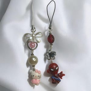 May include: Two phone charms, one with a pink Hello Kitty character and the other with a red Spiderman character. Both charms have silver beads and a silver loop.