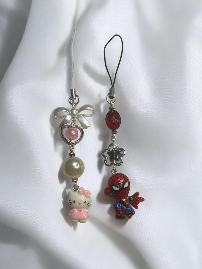 May include: Two phone charms, one with a pink Hello Kitty character and the other with a red Spiderman character. Both charms have silver beads and a silver loop.