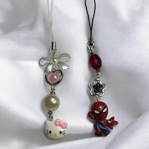 May include: Two phone charms, one with a Hello Kitty character and the other with a Spiderman character. The Hello Kitty charm has a pearl bead, a pink heart, and a silver bow. The Spiderman charm has a red and blue Spiderman figure, a silver star, and a red bead.