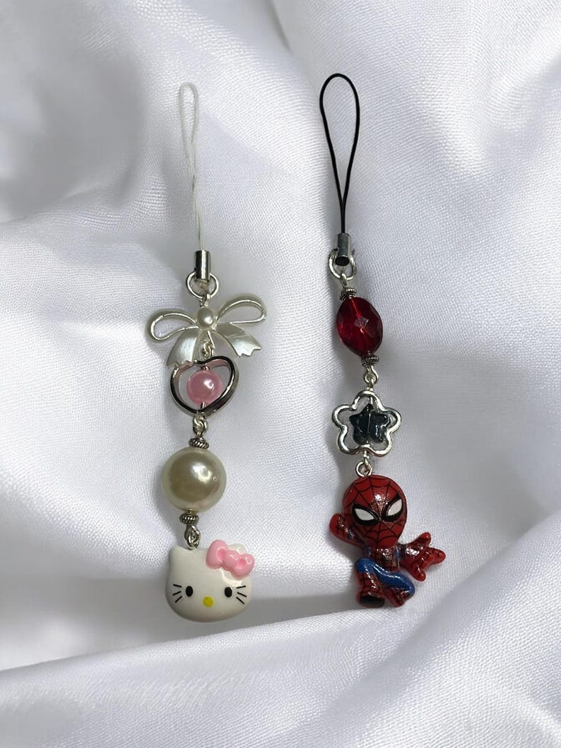 May include: Two phone charms, one with a Hello Kitty character and the other with a Spiderman character. The Hello Kitty charm has a pearl bead, a pink heart, and a silver bow. The Spiderman charm has a red and blue Spiderman figure, a silver star, and a red bead.