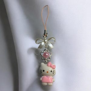 May include: A white Hello Kitty charm with a pink dress and bow, attached to a silver chain with a pink pearl and a white bow. The charm is attached to a pink phone strap.