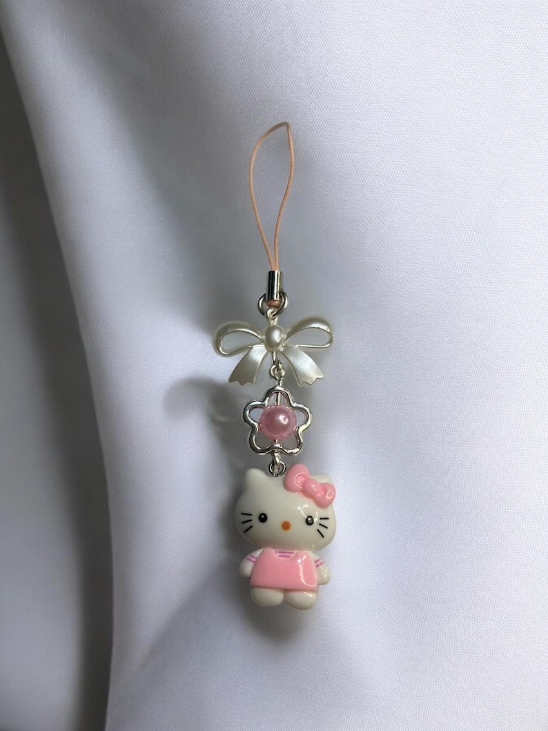 May include: A white Hello Kitty charm with a pink dress and bow, attached to a silver chain with a pink pearl and a white bow. The charm is attached to a pink phone strap.