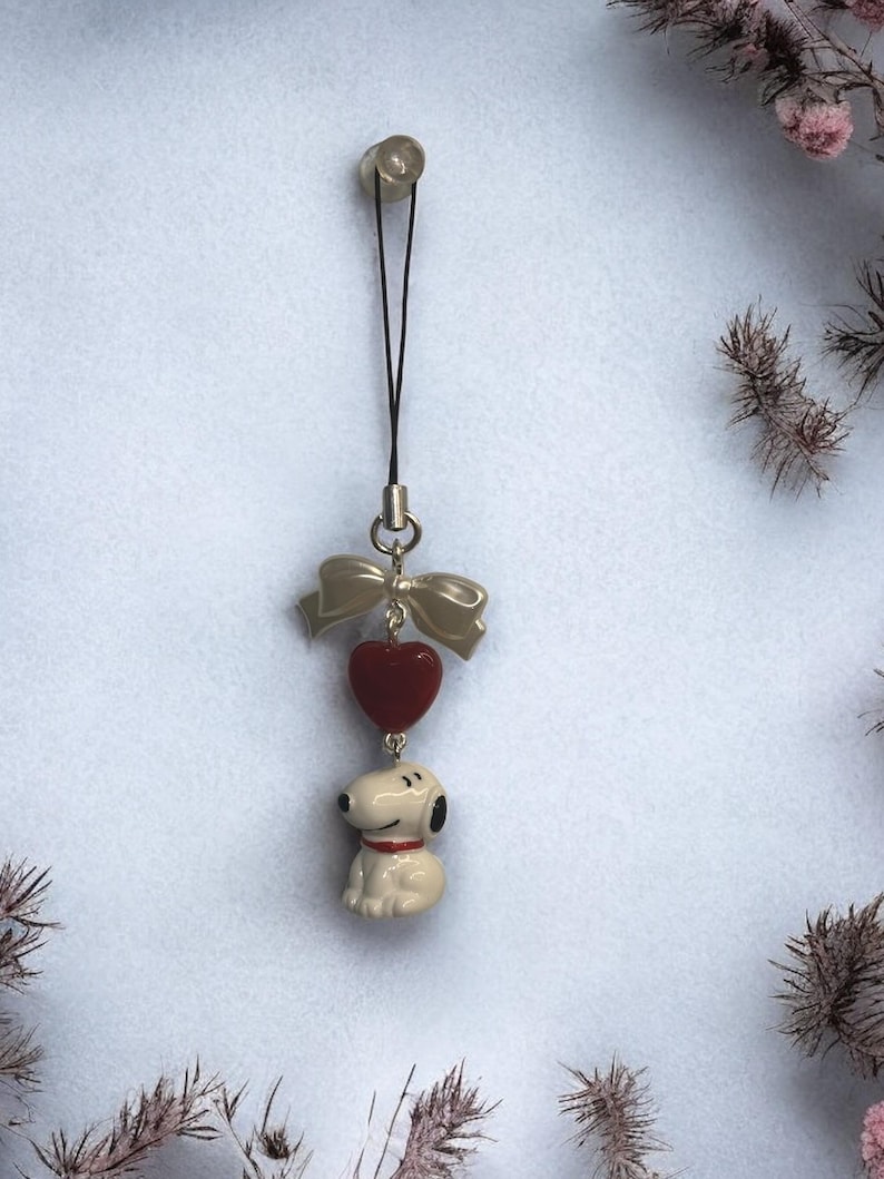 Op de afbeelding: Een wit keramiek beeldje van een cartoonhond met een rode halsband en een rode hartbedel bevestigd aan een zilveren strik. De bedel is bevestigd aan een zwarte telefoonriem.