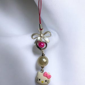May include: A white Hello Kitty charm with a pink bow, a pearl bead, and a silver heart with a pink bead. The charm is attached to a red string.