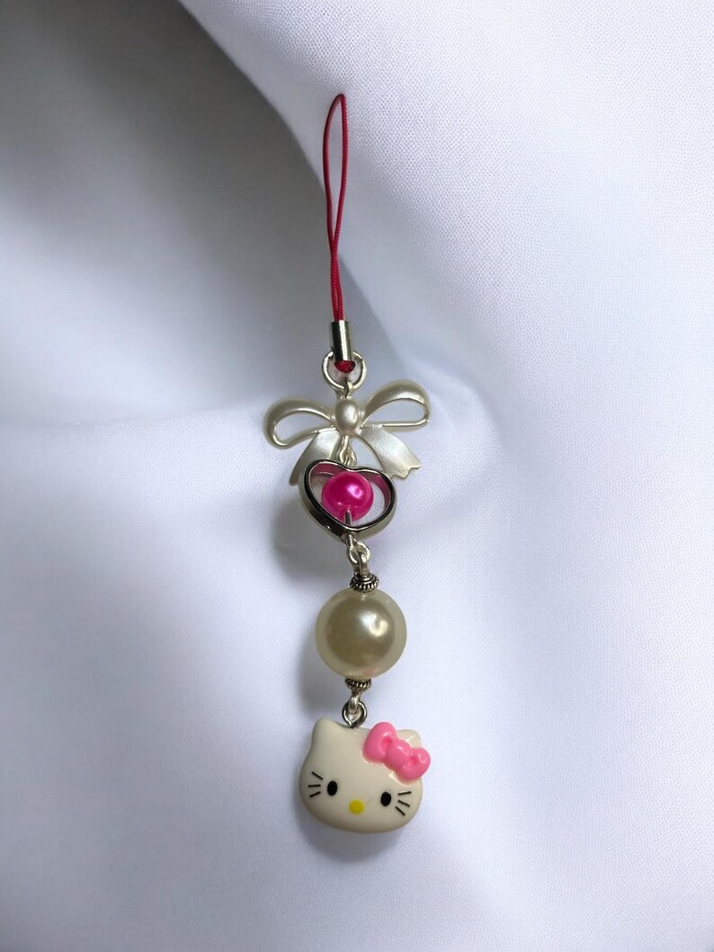 May include: A white Hello Kitty charm with a pink bow, a pearl bead, and a silver heart with a pink bead. The charm is attached to a red string.