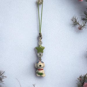 Op de afbeelding: Een groene en witte telefoonbedel met een cartoonhond die een zonnebril en een groen zwempak draagt. De bedel is bevestigd aan een groen touwtje met een doorzichtige plastic kraal aan de bovenkant.