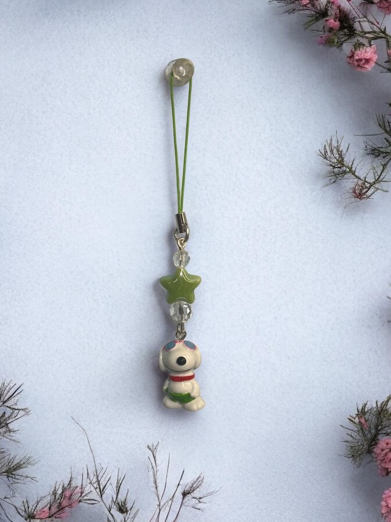 Op de afbeelding: Een groene en witte telefoonbedel met een cartoonhond die een zonnebril en een groen zwempak draagt. De bedel is bevestigd aan een groen touwtje met een doorzichtige plastic kraal aan de bovenkant.