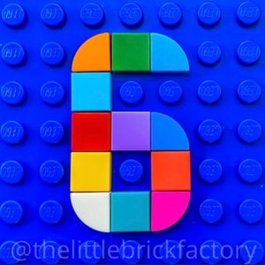 May include: A vibrant number six constructed from Lego bricks, set against a blue Lego base. The number features orange, green, blue, red, yellow, purple, teal, orange, and pink bricks. The base has the text "@thelittlebrickfactory".
