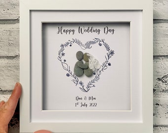 Pode incluir: Uma imagem emoldurada branca com um desenho floral em forma de coração que envolve um casal de arte de seixos. O texto "Happy Wedding Day" está no topo, e "Dan & Mia 1st July 2022" está na parte inferior.