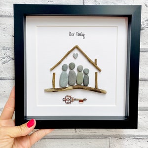May include: A framed pebble art piece depicting a house with a heart above it and five figures standing in front of it. The text "Our Family" is written above the house. A key is placed below the house.