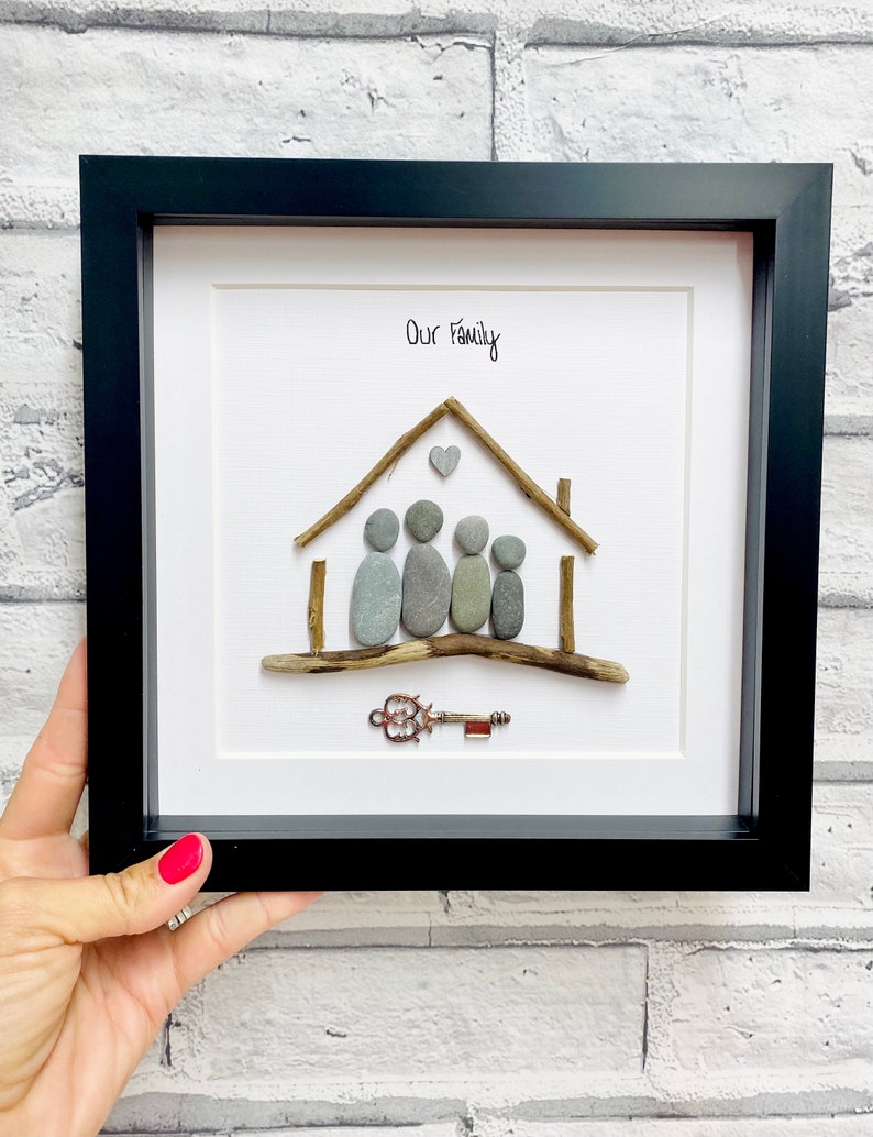 May include: A framed pebble art piece depicting a house with a heart above it and five figures standing in front of it. The text "Our Family" is written above the house. A key is placed below the house.