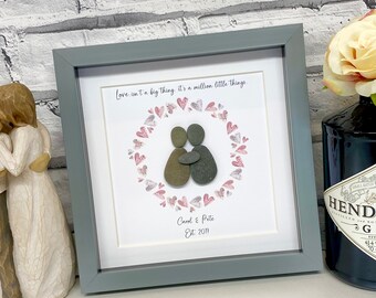 May include: A framed pebble art piece with a couple holding each other in a heart-shaped wreath of pink hearts. The text "Love isn't a big thing it's a million little things." is above the couple. The names "Carol & Pete" and the date "Est. 2011" are below the couple.