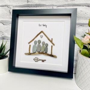 May include: A framed pebble art piece depicting a house with a heart above it and four figures standing in front of it. The text "Our Family" is written above the house. A small silver key is placed below the house.