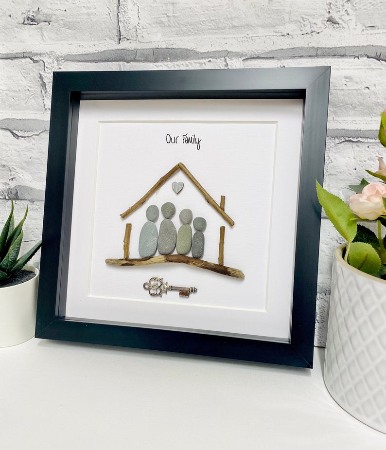 May include: A framed pebble art piece depicting a house with a heart above it and four figures standing in front of it. The text "Our Family" is written above the house. A small silver key is placed below the house.