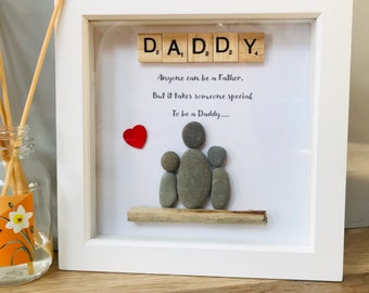 Può includere: Un'opera d'arte in ciottoli incorniciata con una famiglia di tre, un padre e due figli, seduti su un tronco di legno galleggiante. La cornice ha uno sfondo bianco con le parole "DADDY", "Anyone can be a Father, But it takes someone special To be a Daddy..." e un cuore rosso.