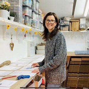 May include: A woman in a gray sweater stands in a workshop, working on a table covered with white paper. She is holding a blue and green heart-shaped object. The table is surrounded by boxes and other supplies.