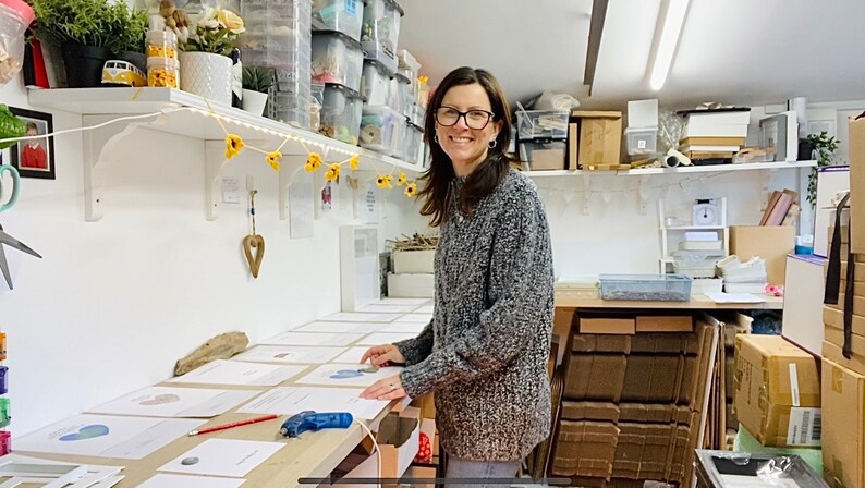 May include: A woman in a gray sweater stands in a workshop, working on a table covered with white paper. She is holding a blue and green heart-shaped object. The table is surrounded by boxes and other supplies.