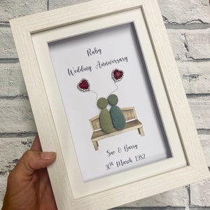 Puede incluir: Una pieza de arte de guijarros enmarcada que representa a una pareja sentada en un banco. El arte se titula "Aniversario de boda de rubí" e incluye los nombres "Sue & Barry" y la fecha "30 de marzo de 1982".
