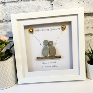 Può includere: Un'immagine incorniciata bianca con un design di arte di ciottoli che rappresenta una coppia che si tiene per mano su un tronco di legno. La coppia tiene due palloncini a forma di cuore dorati. Il testo "Golden Wedding Anniversary" è scritto sopra la coppia. Il testo "Nanny & Grandad The adventures continue" è scritto sotto la coppia.
