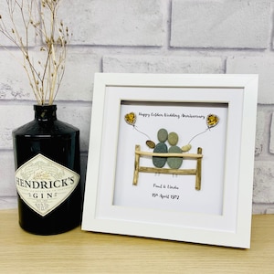 Op de afbeelding: Een ingelijst kiezelkunstwerk ter viering van een gouden huwelijksverjaardag. Het kunstwerk toont twee kiezelstenen figuren op een houten hek, met de tekst "Happy Golden Wedding Anniversary". Links staat een zwarte gin-fles.