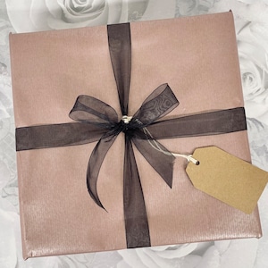 May include: A blush pink gift box with a black ribbon and bow. A brown gift tag is attached to the ribbon.