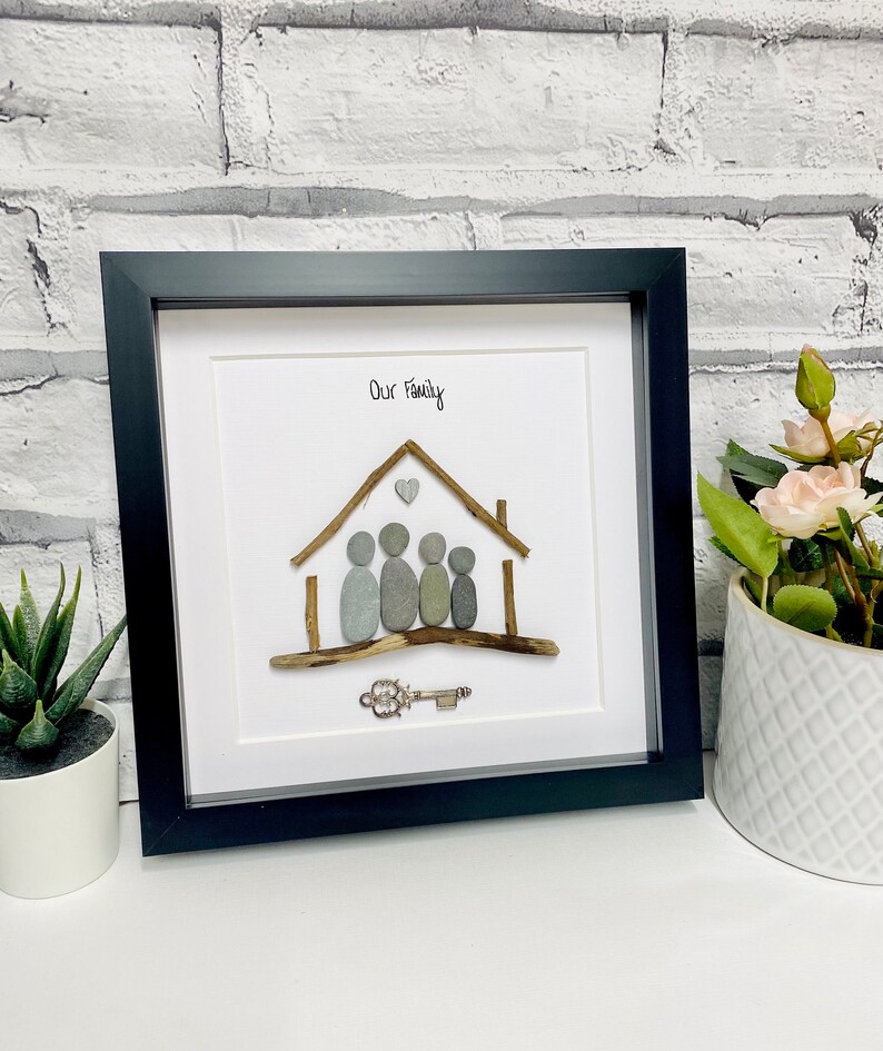 May include: A framed pebble art piece depicting a house with a heart above it and five figures inside. The text "Our Family" is written above the house. A silver key is below the house.