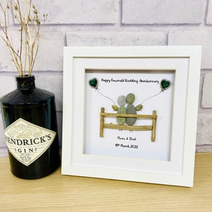 Può includere: Un'opera d'arte con sassi incorniciata in una cornice bianca. L'arte raffigura due figure su una staccionata di legno, con palloncini a forma di cuore verde smeraldo. Il testo recita "Happy Emerald Wedding Anniversary" e "Mum & Dad 19th March 2022". Una bottiglia di gin nera a sinistra.