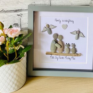 May include: A framed pebble art piece depicting a family of four with two cats. The artwork is on a white background with the words "Family is everything" written at the top. The names "Matt, Lily, Rachel, Mickey, Maisie" are written below the artwork.