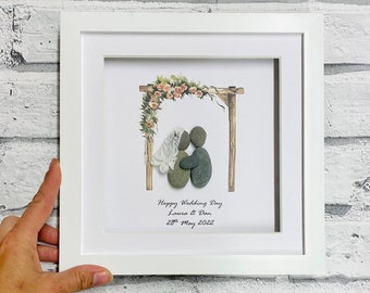 Puede incluir: Una pieza de arte de guijarros enmarcada que representa a una pareja de bodas bajo un arco floral. El texto dice "Happy Wedding Day Laura & Dan 28th May 2022".