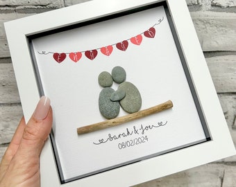 Op de afbeelding: Een ingelijst kunstwerk van kiezelsteen dat een stel toont dat elkaar omhelst op een tak met een banner van harten waarop "Engaged" staat. De datum "08/02/2024" en de tekst "Sarah & You" staan onder het stel.