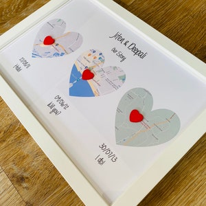 May include: A white framed print with three heart-shaped maps. The maps are labeled with dates and the text "Jiten & Deepali Our Story", "27/08/11 Haselry Ki Hello!", "09/06/12 Will you?", and "30/07/13 I do!"