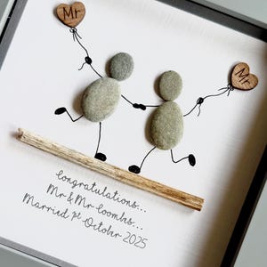 May include: A framed pebble art piece depicting two figures made of pebbles holding hands and walking on a wooden stick. Each figure holds a wooden heart balloon with "Mr" on it. The text reads "congratulations... Mr & Mr looms... Married pt October 2025."